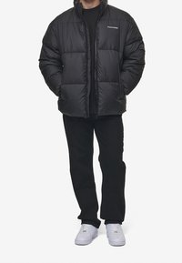 Giubbotto di piumino nero con zip e tasche laterali, caratterizzato da un design trapuntato e dal logo "Peguador" sul petto. Indossato con pantaloni neri e sneaker bianche.