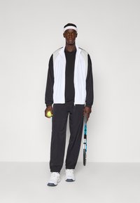 Sort og hvid atletisk outfit med en jakke med lynlås, sort t-shirt, tætsiddende bukser og hvide sneakers. Holder en tennisbold og en ketsjer.