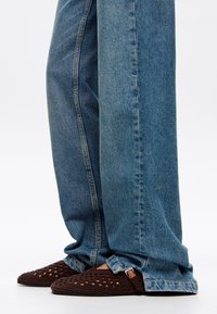 Pantofole all'uncinetto marroni con un design a rete aperta, abbinate a jeans in denim azzurro chiaro a vestibilità morbida e taglio dritto con cuciture visibili.
