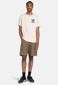 Cremefarbendes Baumwoll-T-Shirt mit navyfarbenem Grafikdesign und Text; kombiniert mit khakifarbenen Shorts und schwarzen Sportschuhen. Einfaches Design, lockere Passform.
