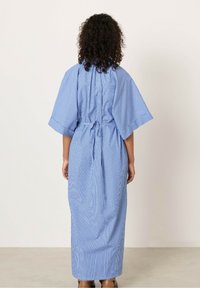 Donna con capelli ricci che indossa un lungo vestito a righe blu e bianche con maniche ampie, legato in vita, che si trova davanti a uno sfondo neutro.