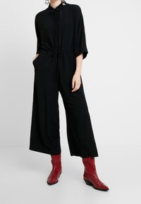 Černý overal s knoflíkovým zapínáním vpředu, krátkými rukávy a šňůrkovým pasem; doplněný o červené kovbojské boty s texturovaným povrchem.