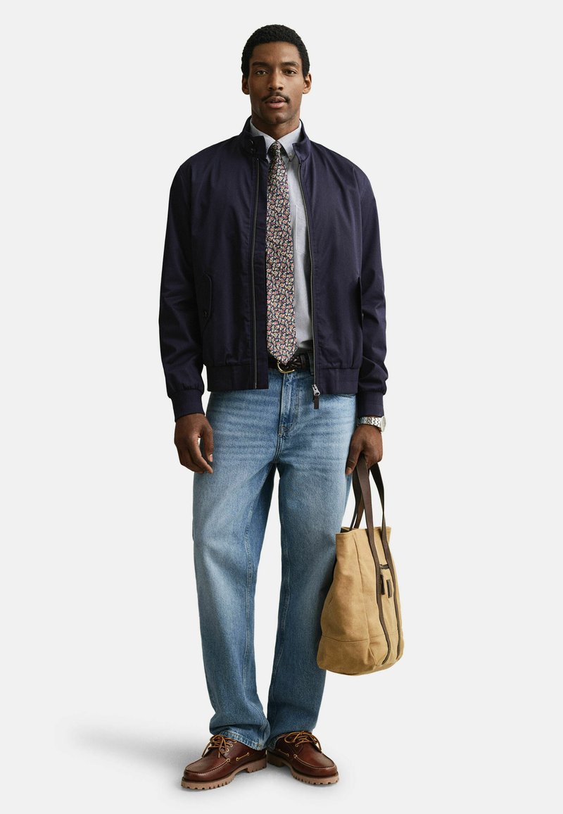Homme portant une veste bleu marine, un jean bleu clair, une cravate à motifs, des chaussures marron, tenant un sac fourre-tout beige à poignées foncées, debout devant un fond blanc.