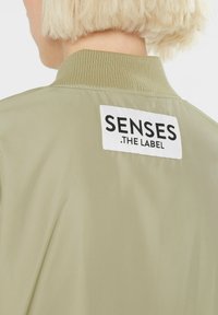 Hellgrüne Bomberjacke mit geripptem Kragen und einem weißen Etikett mit schwarzer Schrift auf der Rückseite. Glatte, glänzende Stofftextur.
