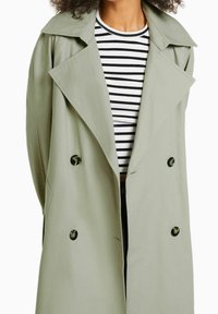 Osoba w jasnozielonym płaszczu trenczowym noszącym czarno-białą koszulę w paski, z rękami za plecami, przycięta na wysokości szyi i w talii.