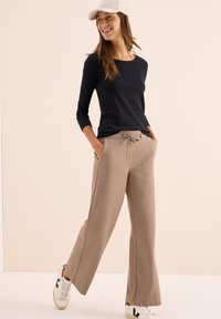 Vrouw die glimlacht, gekleed in beige wijde pijpen broek, zwarte lange mouwen top, witte sneakers en een beige pet, staande tegen een effen lichte achtergrond.
