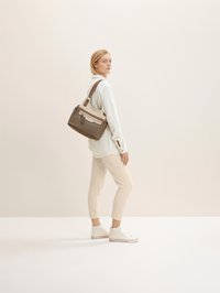 Braune und cremefarbene Handtasche mit einem eleganten Design, ausgestattet mit einer Reißverschlusstasche, glatter Textur und einem Ledergriff. Modell trägt legere Kleidung.