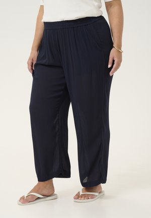 Vrouw die losse donkerblauwe broek, wit topje, gouden armband en witte slippers draagt, staand tegen een effen lichte achtergrond.