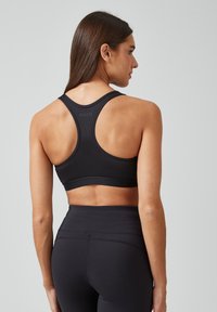 Zwarte sportbeha met racerback-design, gemaakt van een soepele, elastische stof. Heeft een logo op de rug en een ondersteunende band eronder.