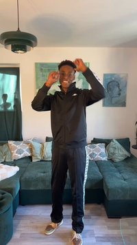 Ung mand i sort træningsdragt smilende og justerer sit hår i en stue med grøn hjørnesofa og vægkunst.