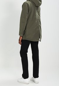 Veste à capuche vert olive à coupe droite, dotée d'ouvertures latérales, d'une texture lisse et de détails en métal argenté. Associée à un pantalon noir et des baskets blanches.