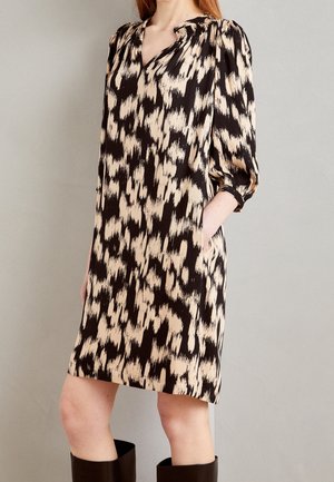 Kobieta ubrana w czarno-beżową sukienkę o abstrakcyjnym wzorze, sięgającą do kolan, z rękawami o długości 3/4 z puffami, oraz czarne kozaki do kolan.