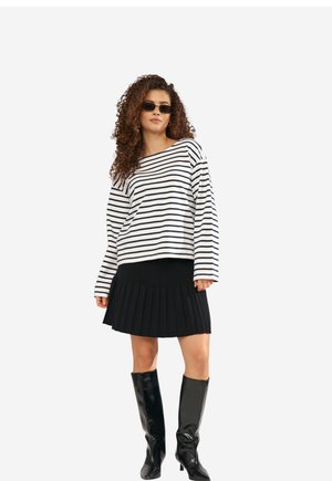 Kvinde med krøllet hår iført sorte solbriller, en hvid og sort stribet langærmet top, sort plisseret nederdel og sorte knæhøje støvler.