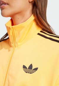 Gelbe Zip-Jacke mit hohem Kragen, verziert mit schwarzen Streifen an den Schultern und einem schwarzen gestickten Logo auf der Brust.