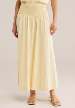 Moteris dėvi šviesiai geltoną, iki kulkšnių siekiančią sijoną su plačia suraukta juosmens juosta ir smėlio spalvos aukštakulnius sandalus su plonais dirželiais.