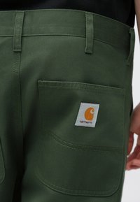 Πράσινο βαμβακερό παντελόνι με τσέπη patch που φέρει πορτοκαλί λογότυπο, ίσια κοπή και ορατές λεπτομέρειες ραφών. Το ύφασμα έχει λεία υφή.