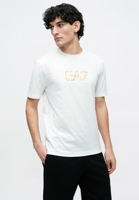 Mladý muž so zvlnenými tmavými vlasmi, oblečený v bielom tričku EA7 Emporio Armani a čiernych nohaviciach, stojaci s jednou rukou v vrecku.