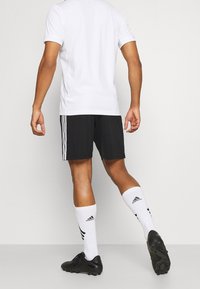 Chemise blanche à manches courtes, shorts noirs avec des accents de rayures blanches, chaussettes blanches avec des rayures noires et chaussures de soccer noires. Vue de dos.