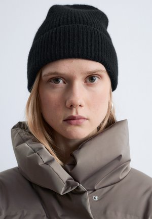 Black ribbed beanie with a snug fit, paired with a light brown padded jacket featuring a high collar and subtle snap closure detail.