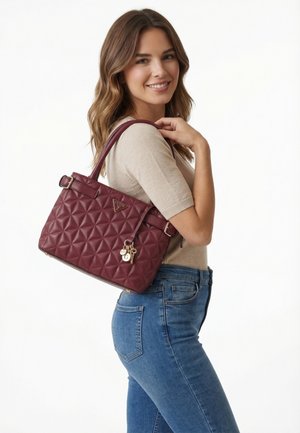 Donna con capelli castani ondulati che indossa una maglietta beige e jeans blu, con una borsa trapuntata bordeaux con un ciondolo dorato sulla spalla.
