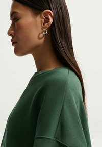 Sweat-shirt vert à coupe décontractée, épaules tombantes et texture douce. Boucle d'oreille argentée visible, avec des cheveux raides et foncés tombant derrière.