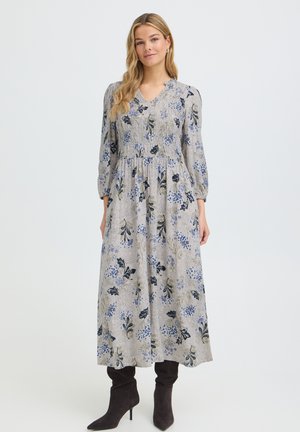 Kvinde med langt blondt hår iført en grå blomsterprintet langærmet midikjole og sorte høje støvler, stående foran en ensfarvet baggrund.