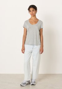 Mlada ženska stoji ob bele stene, oblečena v sivo majico z okroglim izrezom, svetlo oprane ravne kavbojke in sive superge.