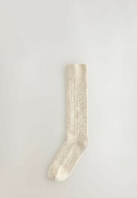 En enkelt beige strikket uldsok liggende fladt p�å en lys neutral baggrund, der viser ribbet struktur og detalje ved kanten.
