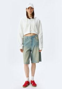 Haut à capuche blanc court, shorts en denim bleu clair, chaussures rouges et chaussettes blanches. Tissu texturé avec une coupe décontractée.