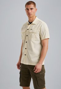 Chemise beige à manches courtes avec deux poches poitrine, boutons noirs et coupe droite, assortie à un short cargo vert foncé avec poches latérales.