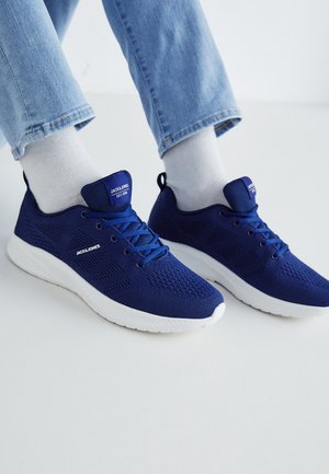 Persona con calcetines blancos y zapatillas tejidas azules con suelas blancas y marca JACK & JONES, combinadas con jeans de mezclilla azul claro.