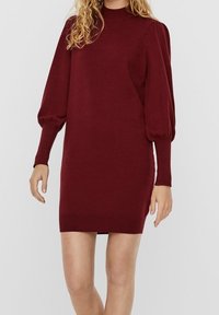 Femme portant une robe en maille bordeaux avec de longues manches bouffantes et un col haut, se tenant devant un fond clair et uni.
