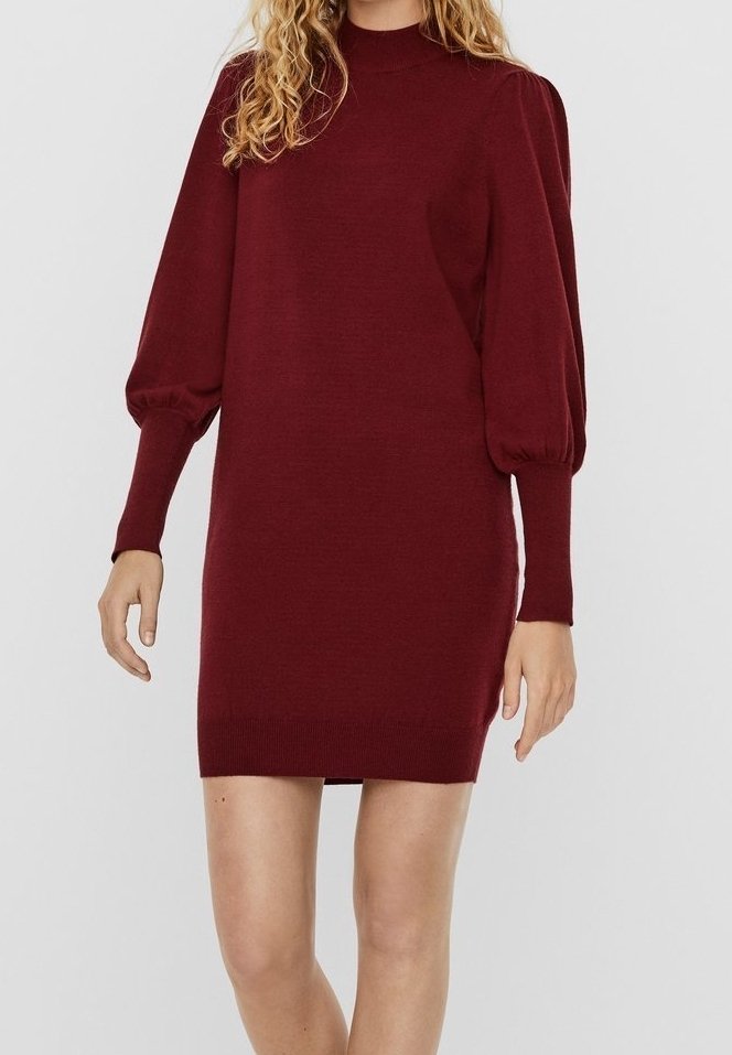 Femme portant une robe en maille bordeaux avec de longues manches bouffantes et un col haut, se tenant devant un fond clair et uni.