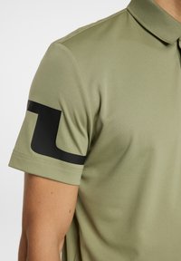 Olivgrön polo med en svart logotyp på vänster ärm, med en mjuk och slät textur samt en klassisk kragsdesign.