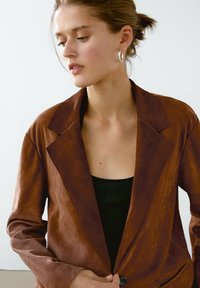 Brun läderblazer med slagkrage och enkel knappstängning. Har en strukturerad yta och långa ärmar. Bärs över en svart topp.