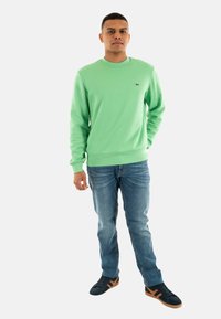 Groene sweatshirt met ronde hals en geribbelde manchetten, gecombineerd met blauwe jeans en zwarte sneakers met bruine accenten.