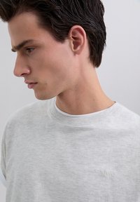 Jonge man met donker haar die een lichtgrijze crewneck sweatshirt draagt met een subtiele "Scalpers"-logo op de borst, met de zijkant naar links gericht in een neutrale setting.