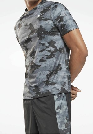 Man draagt een atletisch T-shirt met een grijs-zwart camouflagepatroon en zwarte korte broek met bijpassende camouflagestrips aan de zijkanten, staat met zijn handen achter zijn rug.