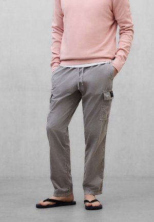 Persona vistiendo pantalones cargo grises, camiseta de manga larga rosa y sandalias negras, de pie con las manos en los bolsillos frente a un fondo liso.