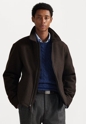 Joven vestido con chaqueta de cremallera marrón oscuro, suéter azul de punto trenzado, camisa de color azul claro con cuello y pantalones grises con cinturón, de pie con las manos en los bolsillos.