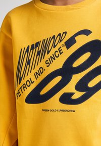 Sudadera amarilla de algodón, con grandes gráficas en azul marino que dicen "NORTHWOOD" y "89", y cuello acanalado.