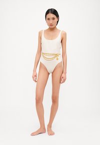 Maillot de bain une pièce blanc avec un décolleté plongeant, mettant en valeur une ceinture en chaîne dorée avec un design superposé et des charms décoratifs. Texture lisse et légèrement brillante.
