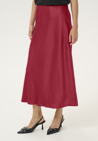 Jupe rouge de longueur mi-mollet faite d'un tissu lisse avec un design fluide. Associée à des escarpins noirs à bride arrière avec des talons aigus et des accents décoratifs.