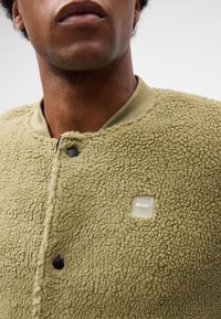 Gros plan sur une personne portant une veste polaire texturée vert olive clair avec des boutons pression et un petit écusson logo "EST. 1993".