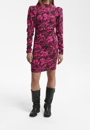 Femme portant une robe ajustée à manches longues rose et noire à motifs avec des épaules bouffantes, associée à des bottes noires montant jusqu'aux genoux.