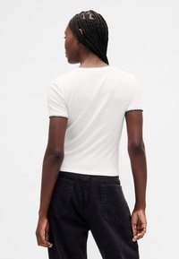 Hvit kortermet topp med kontrasterende svart kant ved ermene og halsen. Jevn tekstur og tettsittende design. Svarte bukser synlige nedenfor.