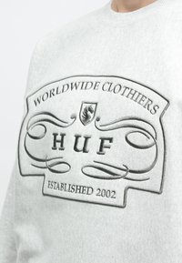 Ανοιχτό γκρι φούτερ με έντονο κεντημένο λογότυπο που αναγράφει "HUF" και "WORLDWIDE CLOTHIERS ESTABLISHED 2002."