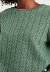 Grüner strukturierter Pullover mit Zopfmuster, rundem Halsausschnitt und weichem Stoff. Das Design zeigt erhabene, blattähnliche Motive.