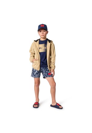 Beige lichtgewicht jack met zwarte bies, marineblauw T-shirt met gouden logo, navy shorts met patroon en marineblauwe/rode sandalen. Eenvoudige, casual outfit.