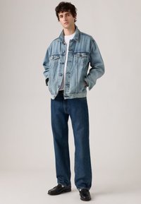 Denim jacket in light blue, featuring two front chest pockets, button closure, and relaxed fit, paired with dark blue jeans and black shoes.